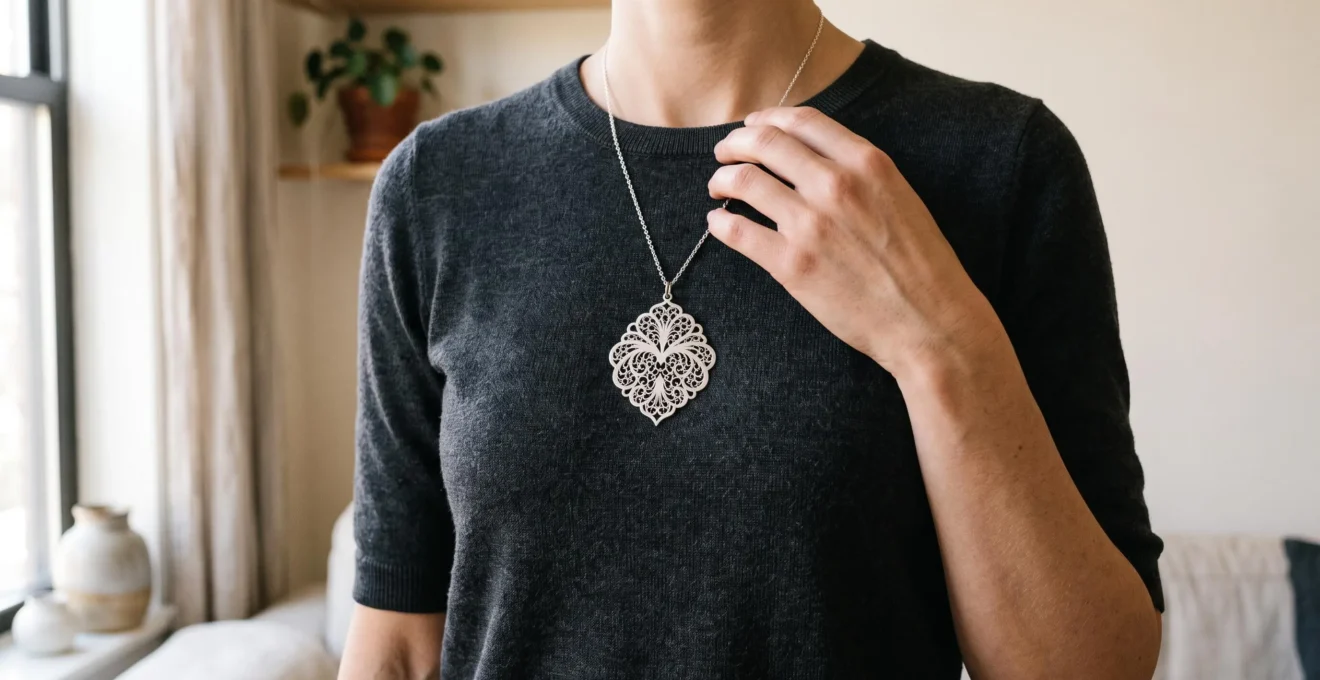 A woman in a plain charcoal top adjusts a matte silver filigree pendant resting at her collarbone in soft daylight.
