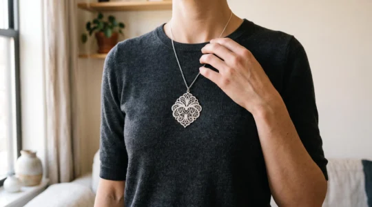 A woman in a plain charcoal top adjusts a matte silver filigree pendant resting at her collarbone in soft daylight.